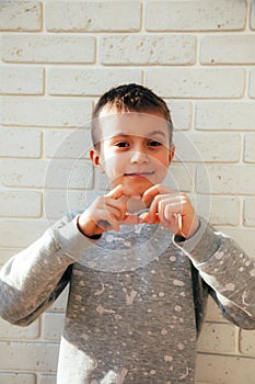 The boy smiles and shows a heart with his fingers. Portrait of a child against a white brick wall