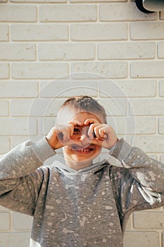 The boy smiles and shows a heart with his fingers. Portrait of a child against a white brick wall. Close up