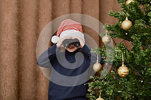 The boy smiles and watches gifts near the Christmas tree, looks out through binoculars. Dressed in a carnivalcap