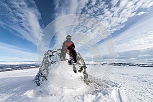 Boy in snowmobile driver clothes sits on the big stone point marking Swedish Norwegian state border in Lappland.