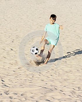 Boy soccer player kicks football ball on beach on sea sand. Beach soccer, summer holidays, sport games outdoor concept