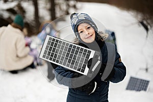 Boy with solar panel battery at hand against his family in winter forest spending time together on a picnic