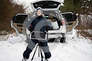 Boy with solar panel battery on tripod against car in winter woods