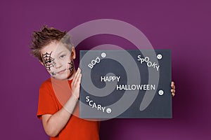 A boy with a spider and spider web drawn on his face, holding a blackboard with the text of Halloween