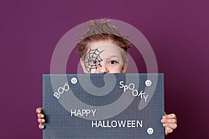 A boy with a spider web drawn on his face, holding a blackboard with the text of Halloween