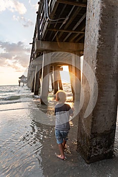 Boy standing under the pier in water watching sunset through concrete supports