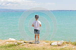 Boy stands on coast of lake, sunny daytime. Healthy leisure. Summer. Calmness. Nature. Panoramic view. Blue clear water