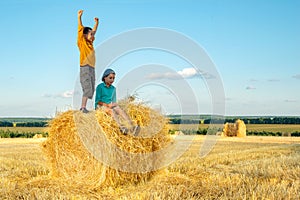 The boy stands on a haystack and joyfully raises his hands up to the sun
