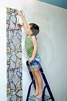 A boy stands on a ladder and glues wallpaper