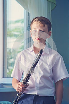 The boy stands near the window and holds a black clarinet in his hands, looks into the camera. Musicology, music education and edu