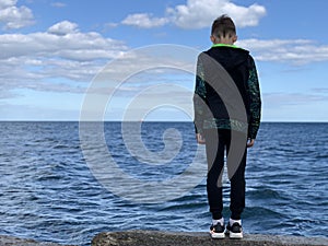 The boy stands on the shore facing the sea. A child on the pier looks at the sea. A teenager by the ocean