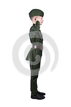 Boy stands sideways in military uniform greets, puts his hand to his head isolated on white