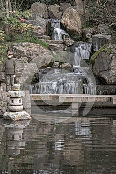 A boy stands on a stone bridge in front of waterfall and rocky cliffs in Kyoto garden