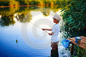 Boy in straw hat with a fishing rod catches fish on riverbank at golden hour of sunset, river