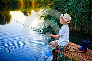 Boy in straw hat with a fishing rod catches fish on riverbank at golden hour of sunset, river