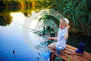 Boy in straw hat with a fishing rod catches fish on riverbank at golden hour of sunset, river