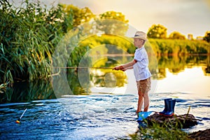 Boy in straw hat with a fishing rod catches fish on riverbank at golden hour of sunset, river