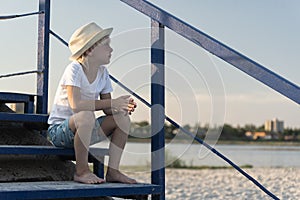 Boy in straw hat sitting on stairs in beach background. Summer vacation