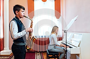 Boy student learns to play the saxophone in a music lesson to accompaniment of the piano