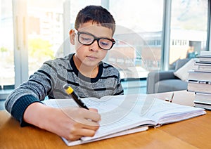 Boy, student and writing in book for studying, homework or learning in education on table at home. Smart little kid or