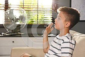 Boy suffering from heat near fan at home. Summer season