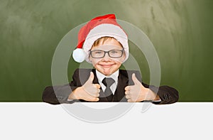 Boy in a suit with red christmas hat looks out from behind a banner and showing thumbs up