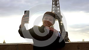 Boy in a suit is taking a selfie on phone on the background of the Eiffel tower
