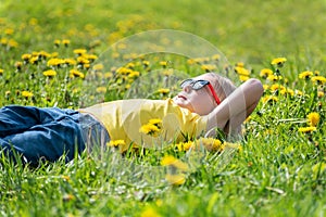 Boy in sunglasses lying down in the green grass. Child rest in dandelions flower filed. Sunny day