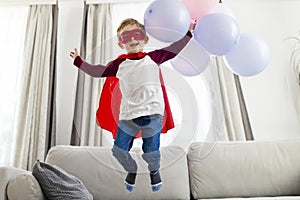 Boy in superhero costume with balloons jumping on sofa