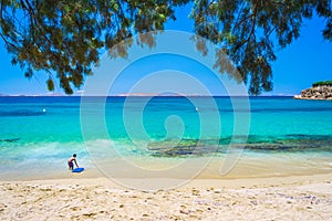 Boy with surf board at the tropical beach of Agios Stefanos with tamarisk and turquoise water, Mykonos island, Greece