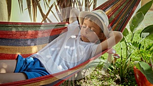 A boy sways and rests in a hammock at garden, portraying the sensations of summertime, treasured childhood memories, and