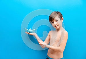 A boy in swimming trunks holds a paper boat on a blue background. Background travelers, sea adventures. Fun at the water park and