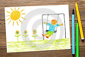 A boy swings on a swing at noon. Children's drawing with felt-tip pens. Wooden table