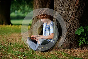 The boy with the tablet in hands sits under a big tree.