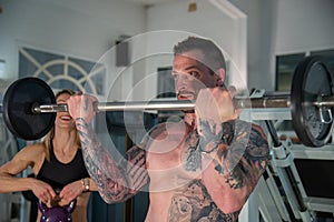 Boy with tattooed body does workout in the gym using weights, girl with kettlebell.