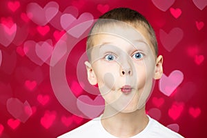 Boy teenager 7-10 in white t-shirt makes faces depicting kiss, with wide open eyes on red and pink hearts bokeh
