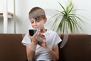 Boy teenager holds smartphone in his hands and smiles. Child at home on couch with phone in his hands