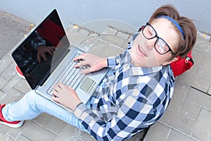 Boy teenager schoolboy or student is sitting on the stairs, working in the computer, wearing glasses, in a shirt, smiling, red b