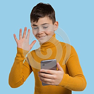 Boy teenager in a yellow sweater, with a smartphone in hand, onine communication, welcomes the interlocutor, smiling