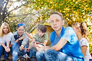 Boy and teens sit on campsite with sausages