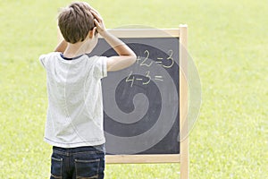 Boy thinking, writing and counting on blackboard. Green outdoor background
