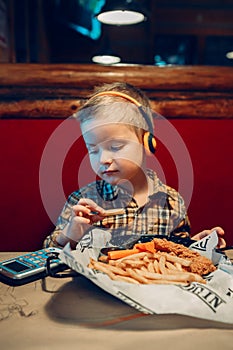 boy toddler playing digital toy cell phone gadget with ear phones and eating food meal in restaurant