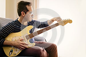 Boy tuning his electric guitar sitting on the sofa at home during confinement