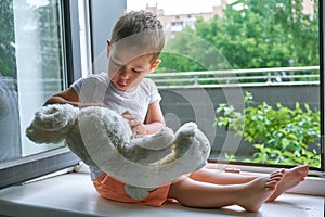 Boy of two years sitting by the window and hugs a toy Bunny. rainy weather, waiting for dad to come home from work