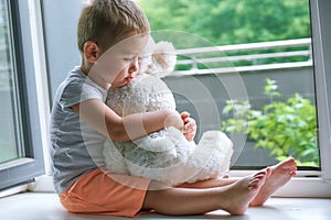 Boy of two years sitting by the window and hugs a toy Bunny. rainy weather, waiting for dad to come home from work