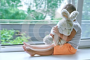 Boy of two years sitting by the window and hugs a toy Bunny. rainy weather, waiting for dad to come home from work