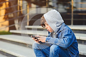 Boy use phone and plays games. Kid in the hood sitting on the stairs. Kids addicted online games and cartoons. Schoolboy plays on