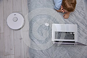 Boy uses a laptop on the bed while the robot vacuum cleaner does the cleaning