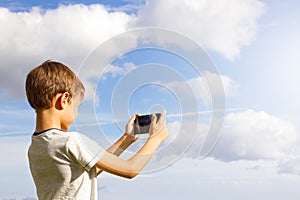 Boy using mobile phone. Child taking photo with his smartphone. Beautiful sky background. Back view. Technology concept