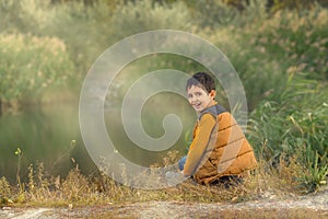 A boy in a vest sits on the shore of a lake